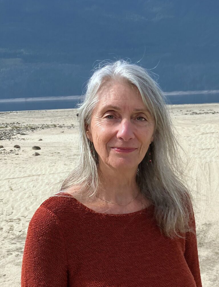 A photo of Deidre Simon Dore. She is a light skin toned woman with long grey hear, standing on a beach with the water in the background.