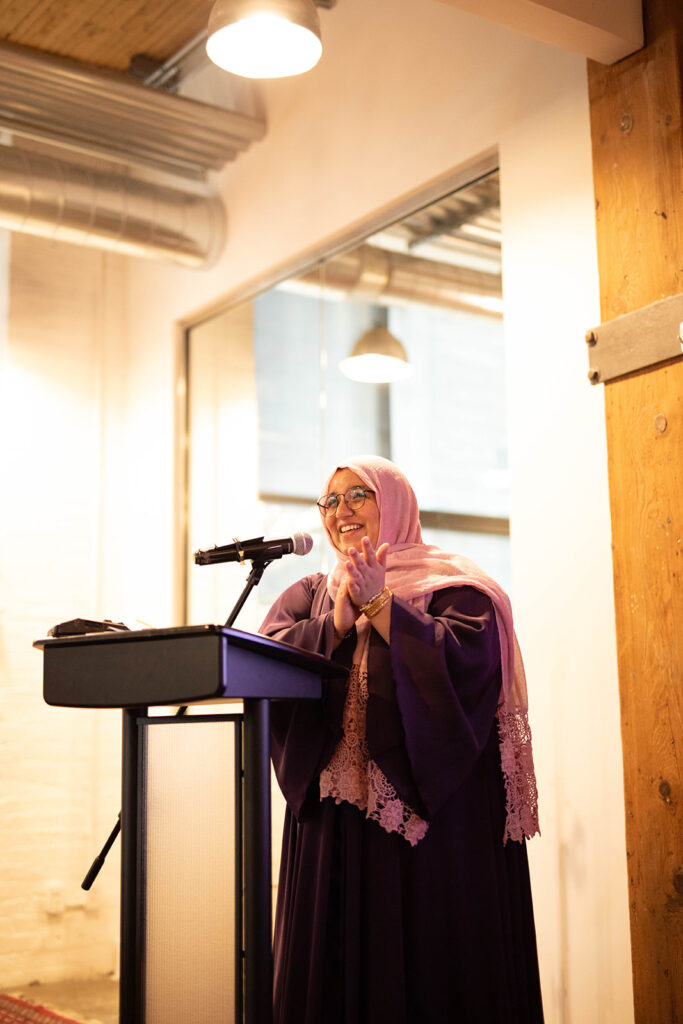 A photo of Narjis Sheikh at the podium at the Muslim Literary Festival. She is wearing a pink headscarf and beams in excitement at the audience, clasping her hands together. 