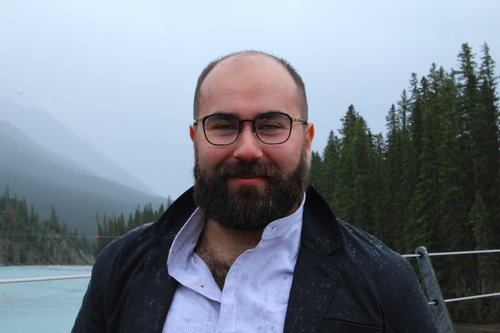 A photo of writer Ben Berman Ghan. He is a bald, light skin toned man with a full, dark beard and glasses. He stands before a lake, pine forest, and foggy mountains in the distance.