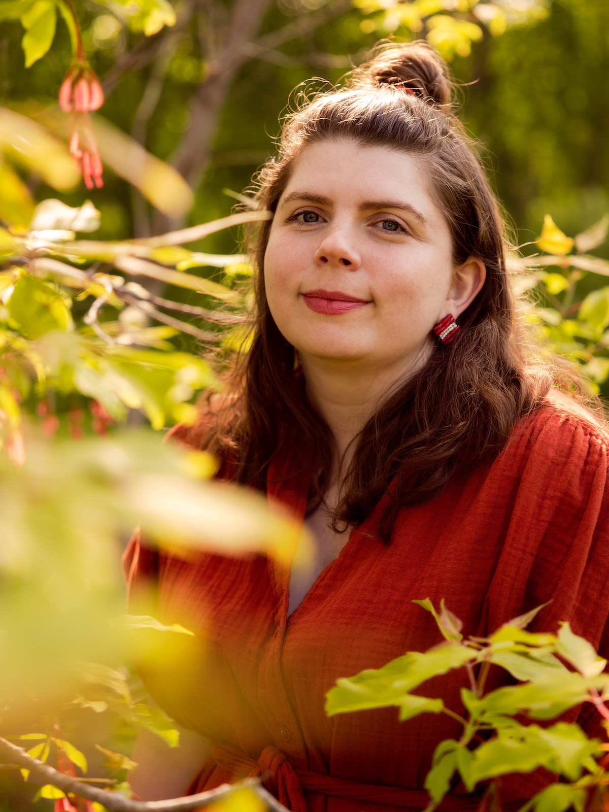 A photo of writer Jess Taylor. She is a light skin-toned woman with dark, medium length hair, half pulled into a topknot. She wears red lipstick and a red shirt, and stands among green foliage in golden hour.