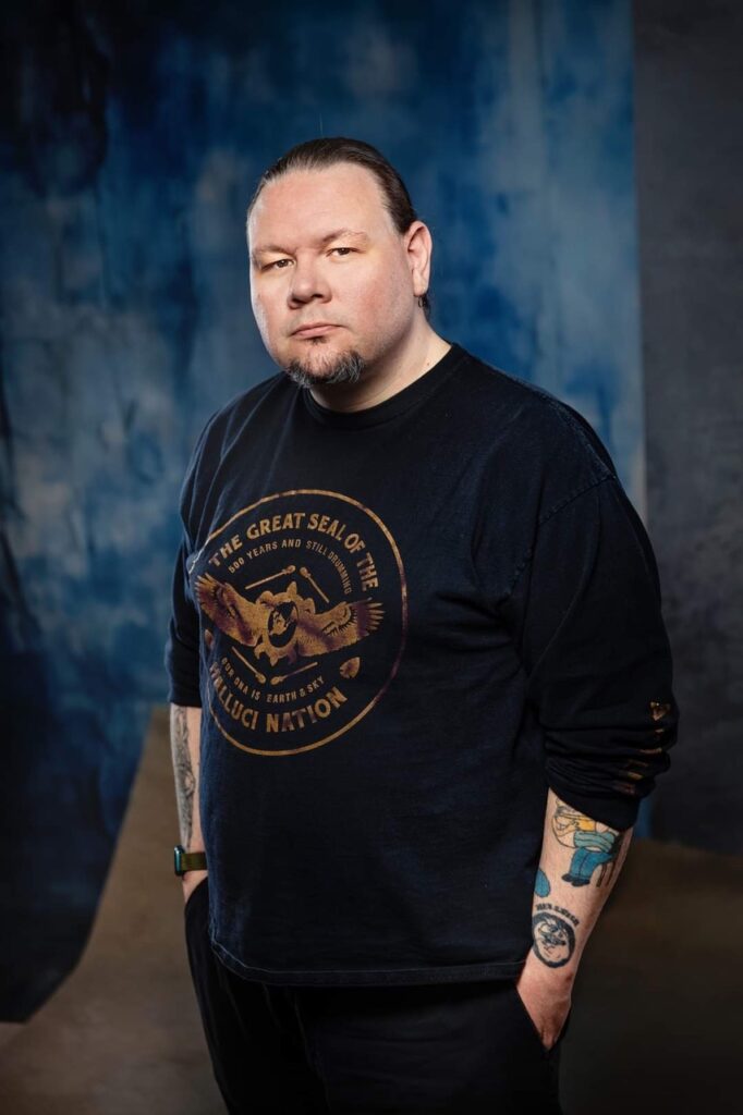 A photo of Nehiyawak-Metis writer John Brady McDonald. He is a man with hair tied in a ponytail and a goatee, wearing a black shirt with the sleeves pushed up to reveal tattoos along his arms.