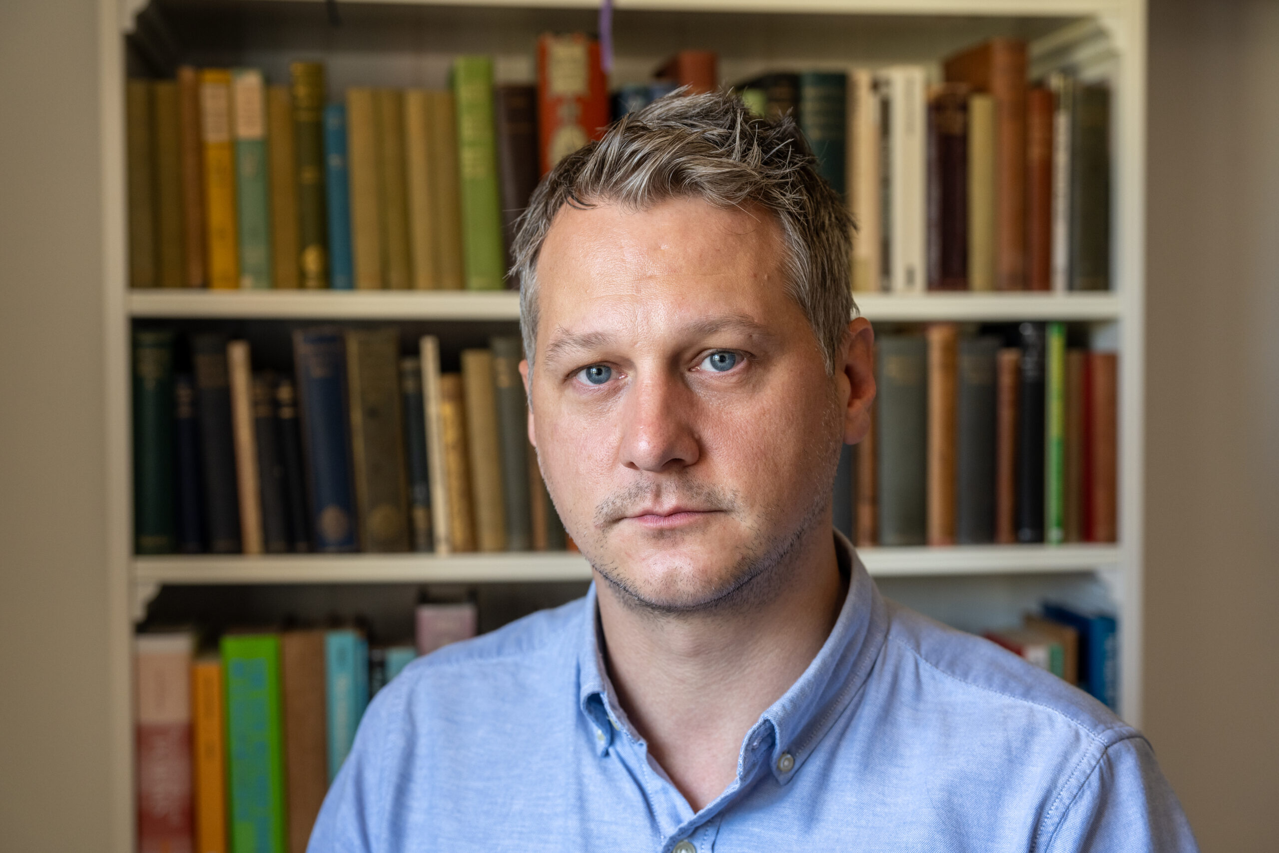 A photo of writer Adrian Markle. He is a light skin-toned man with short, greying hair, wearing a blue oxford shirt and standing in front of a bookcase.