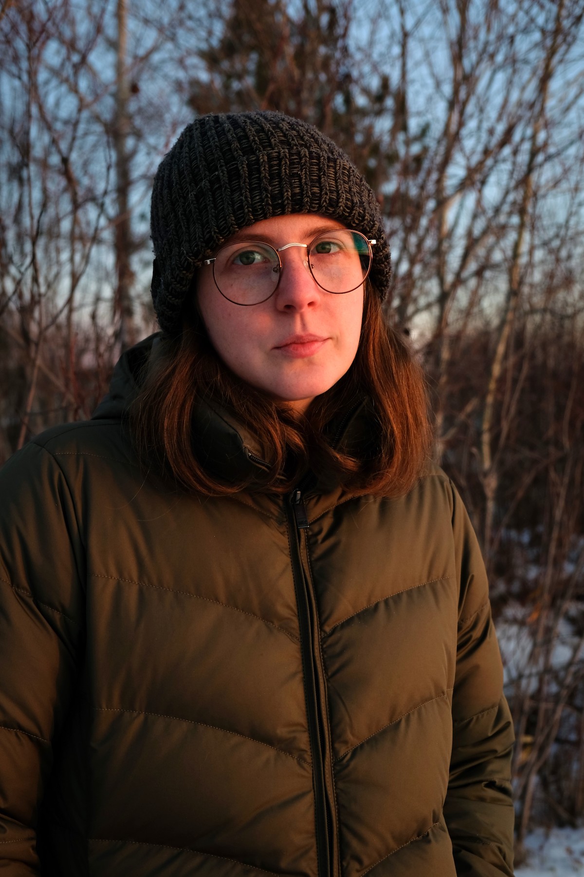 A photo of poet Joelle Barron. They are a light skin-toned person with shoulder-length brown hair and large glasses, and wear a tuque and puffer jacket outside, at dusk, the bare branches of trees behind them.