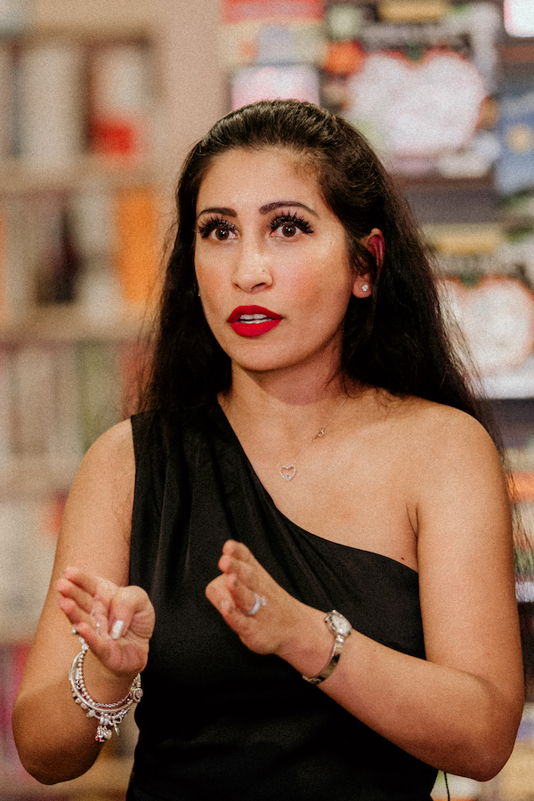 A photo of writer Taslim Burkowicz. She is a medium skin toned woman with long, dark hair and red lipstick. She wears a one-shoulder black top and is gesturing.