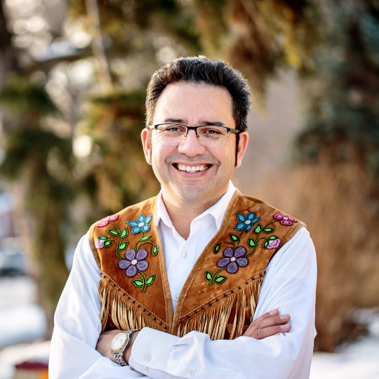 A photo of writer Richard Van Camp. He is a Tlicho Dene man with short, dark hair and glasses, and a big smile. He wears a beautiful vest made of hide with beaded flowers on it and stands outside, with trees in the background.