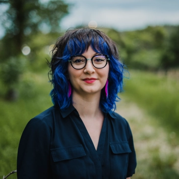 A photo of writer Courtney Bates-Hardy. She is a light skin-toned woman with shaggy-cut blue/purple hair, red lipstick, and glasses. She stands outside in a grassy field.