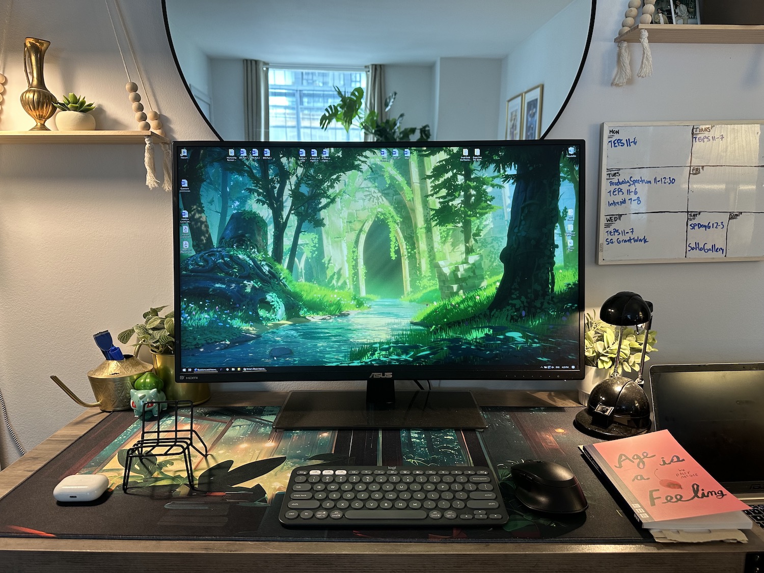 A photo of Makram Ayache's workspace, showing a large monitor with an illustration of a peaceful, abandoned temple in a woodland, a copy of Age is a Feeling, and a whiteboard calendar on the wall. A round mirror behind the monitor reflects a window and large monstera plant.