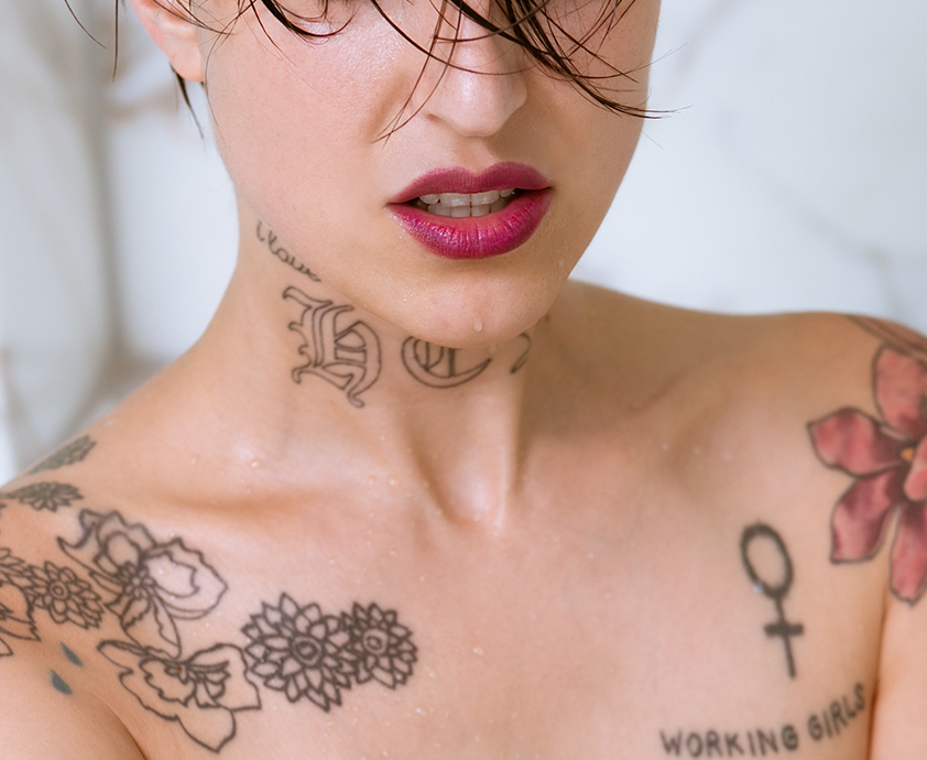A photo of Angel B.H., cropped to hide her eyes from view. She is a light skin-toned woman with tattoos on her neck, bare shoulders, and chest. She wears red lipstick and has a slight sneering expression.