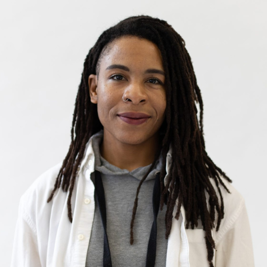 A photo of writer Val Bah. They are a nonbinary Black person with long dreadlocs, wearing a white shirt over a grey hoodie, smiling at the camera.