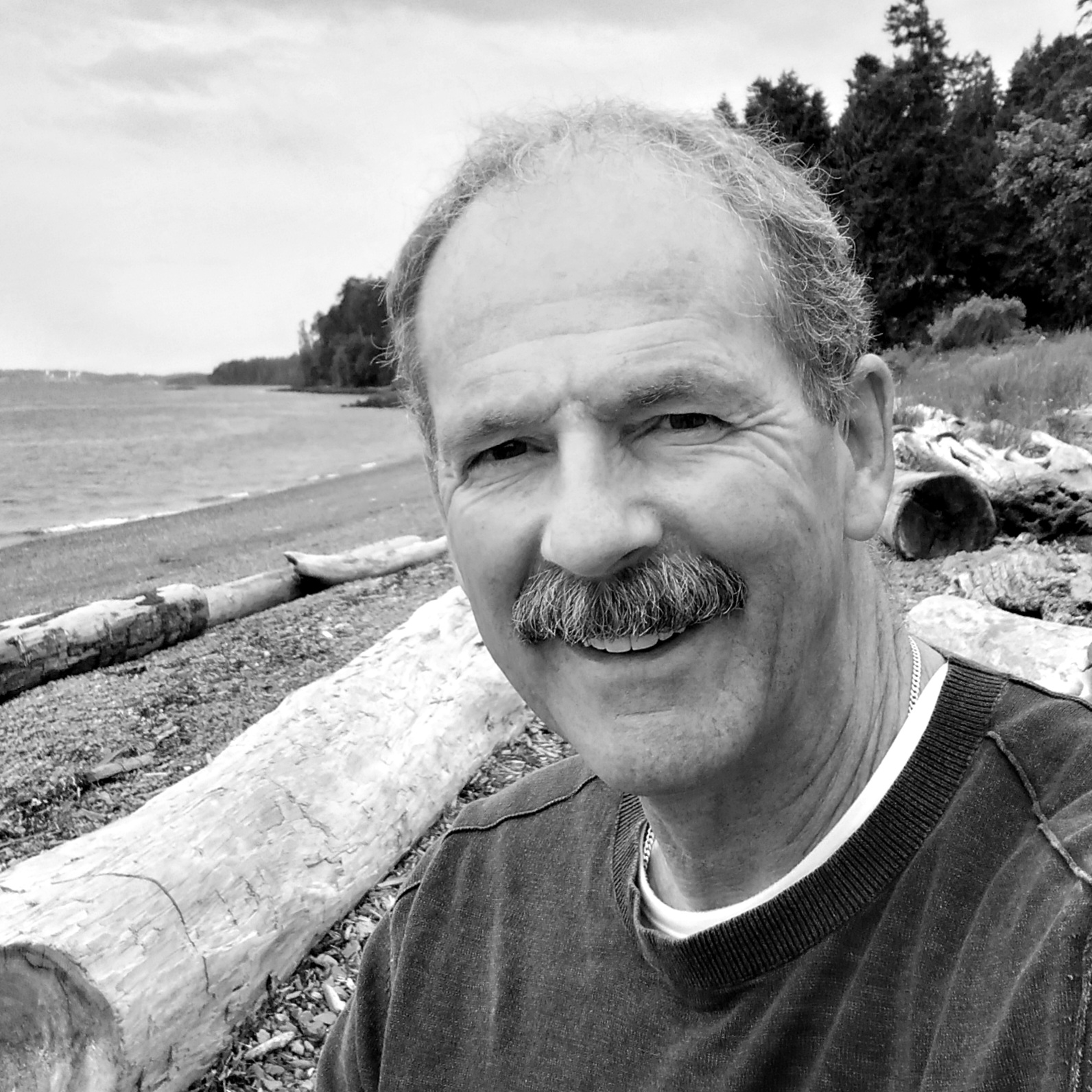 A black-and-white photo of writer Danial Neil. He is a light skin-toned man with light coloured hair and a bushy moustache, sitting on a log on a beach, with the waves lapping the shoreline and dark, coniferous trees in the background.