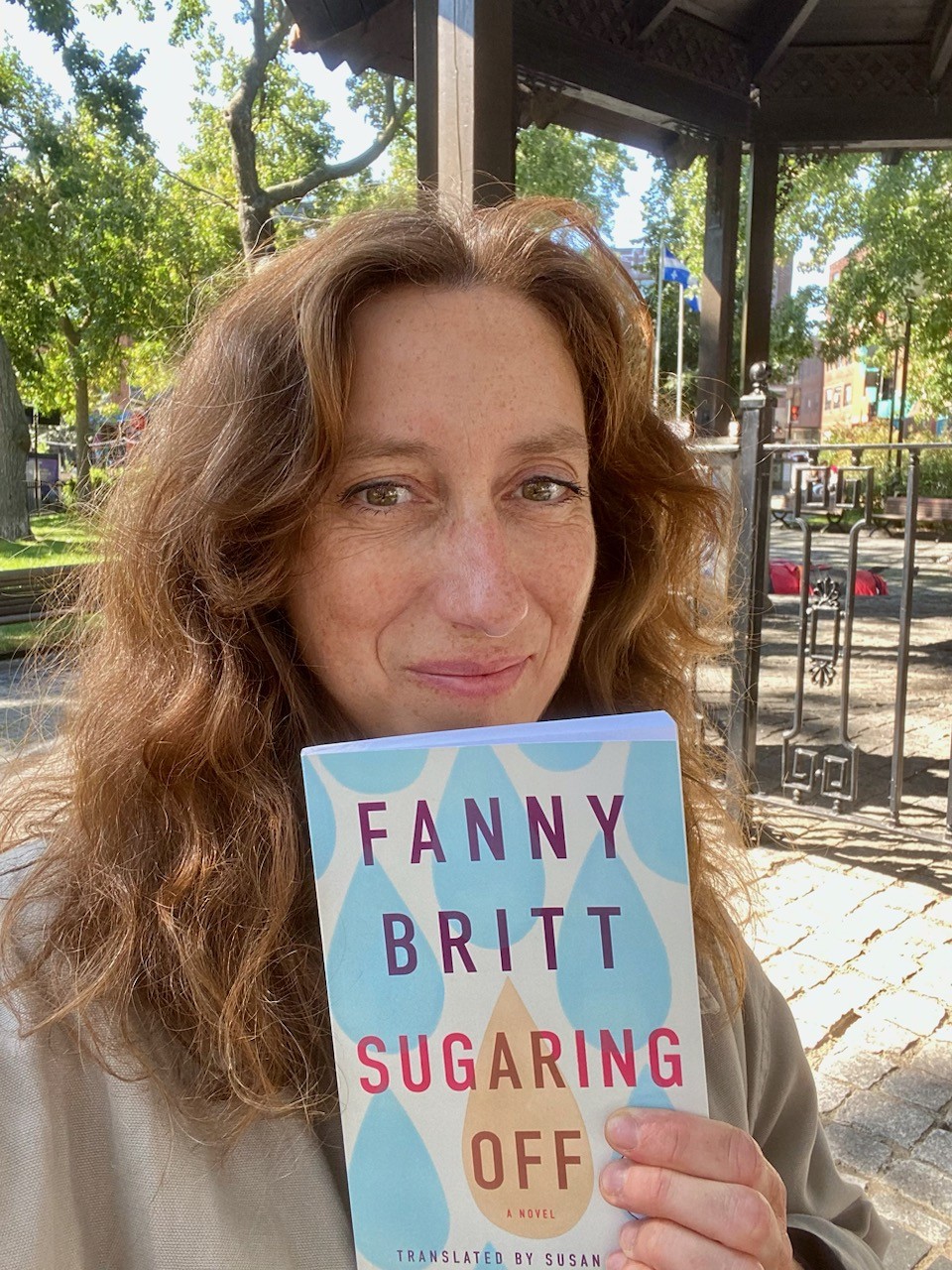 A photo of author Fanny Britt, a light skin-toned woman with medium brown, wavy hair, holding a copy of her book Sugaring Off outdoors.