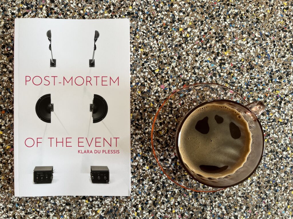 An aerial view of the book Post-Mortem of an Event by Klara du Plessis beside a cup of coffee.