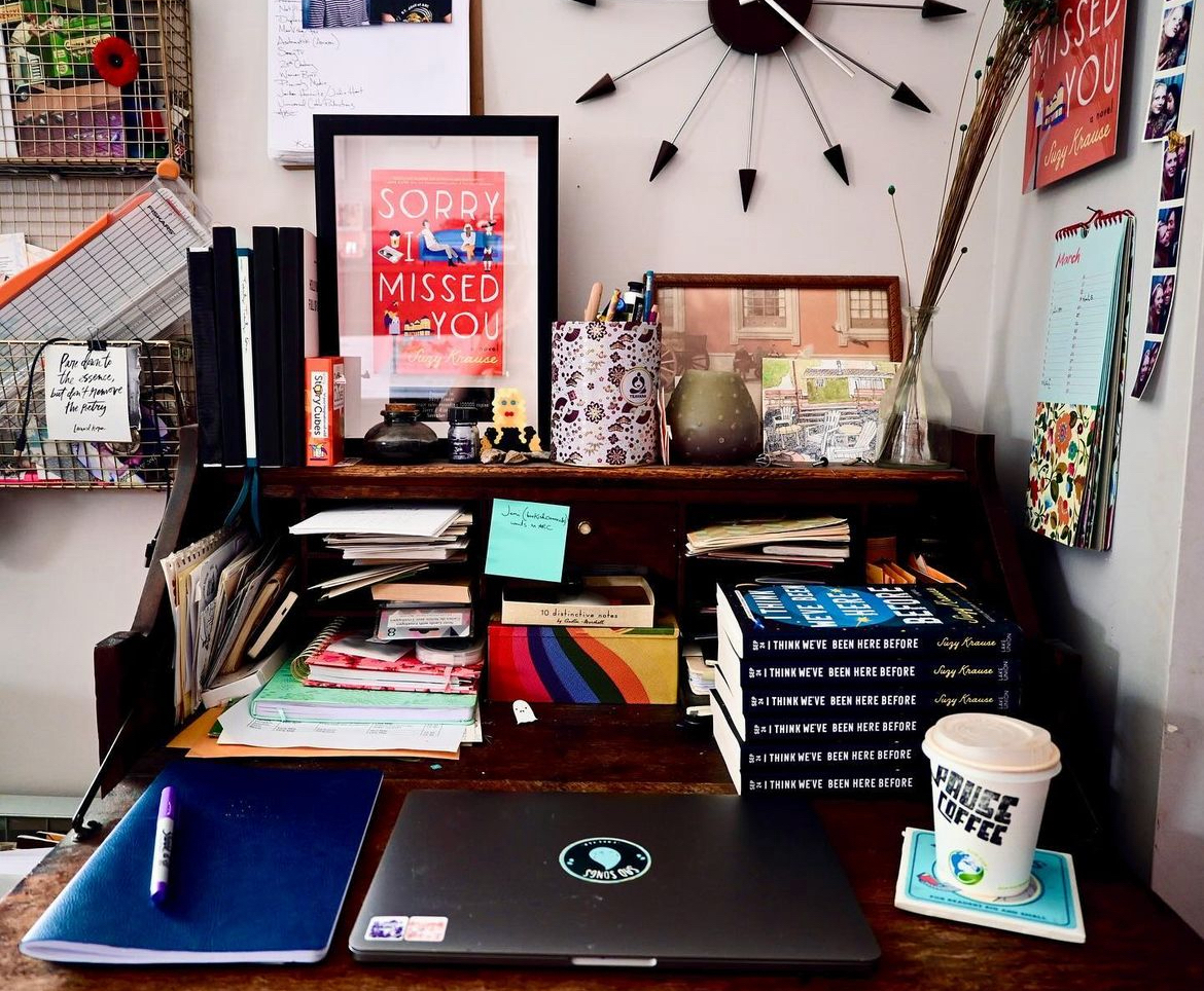 A photo of Suzy Krause's workspace, featuring a stack of her novels, framed photos, a vintage clock, a closed laptop, and a takeaway coffee cup.