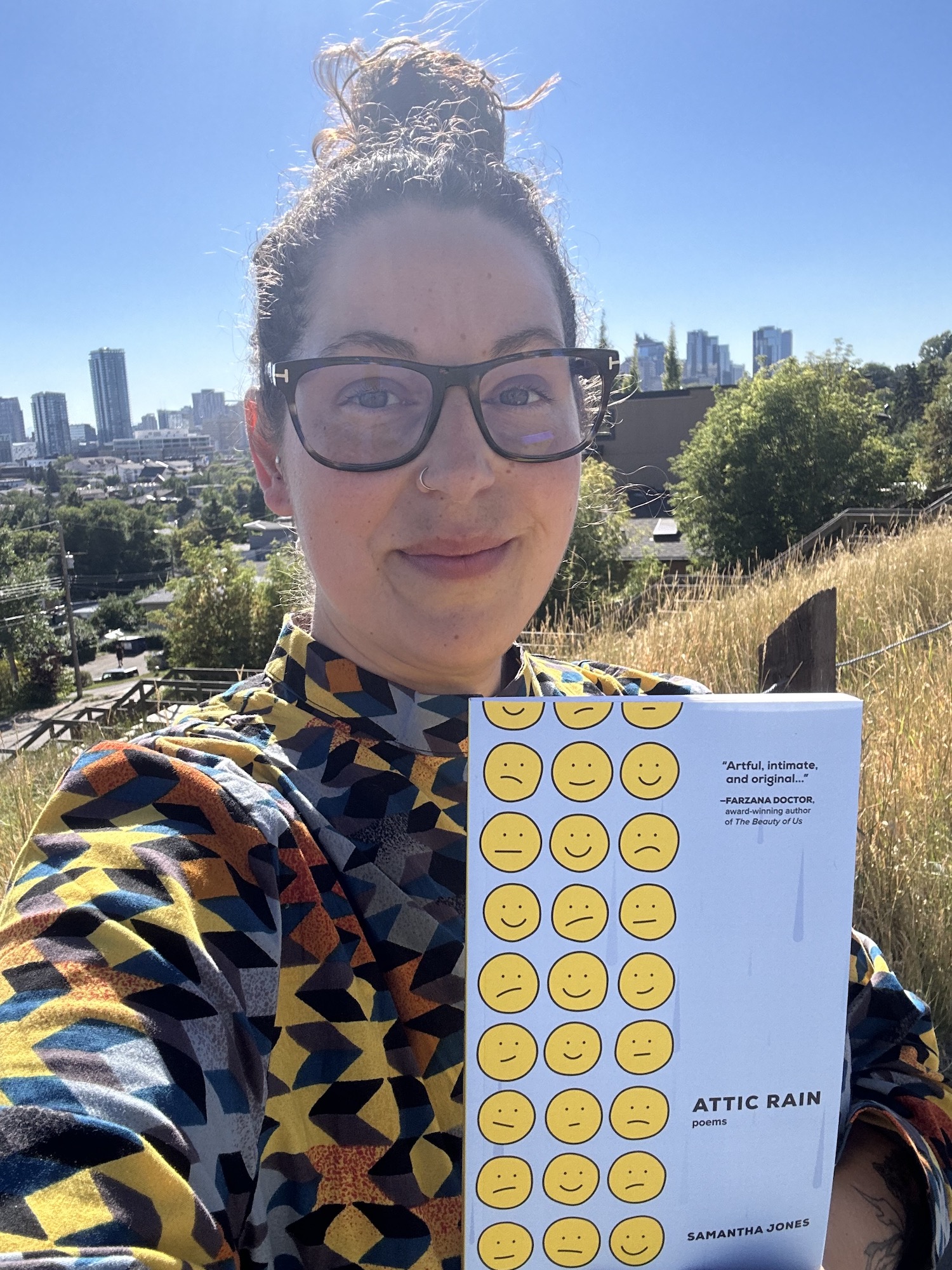 A selfie photo of Samantha Jones holding her book Attic Rain outside on a sunny day in Calgary, with the skyline in the distance.