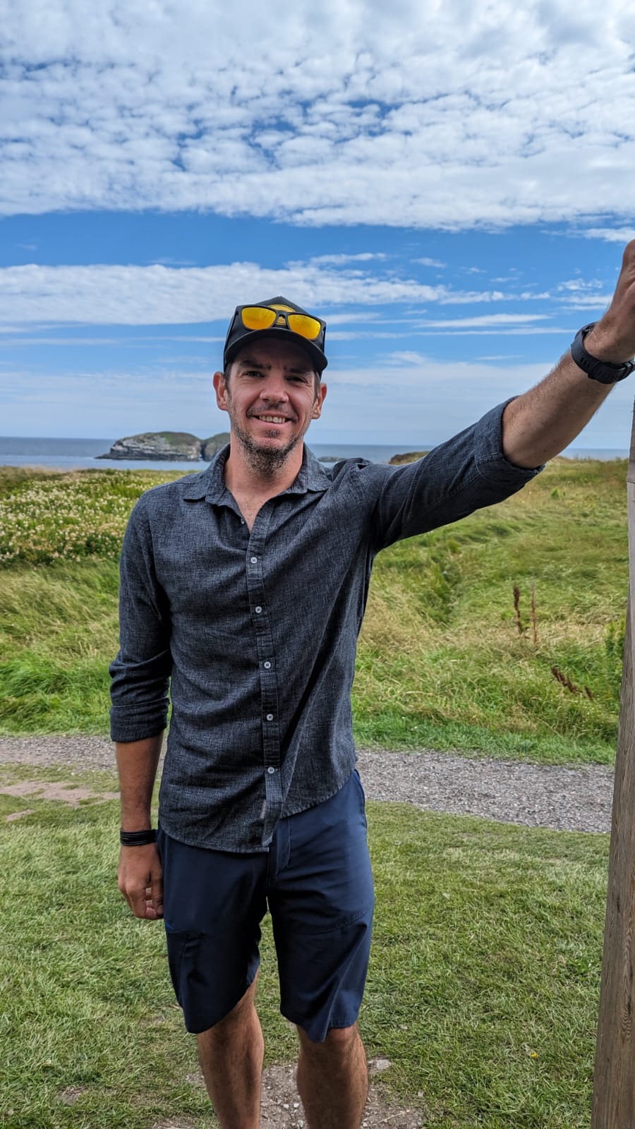 A photo of Douglas Walbourne-Gough. He stands on a grassy cliffside overlooking a giant rock island in the sea. 
