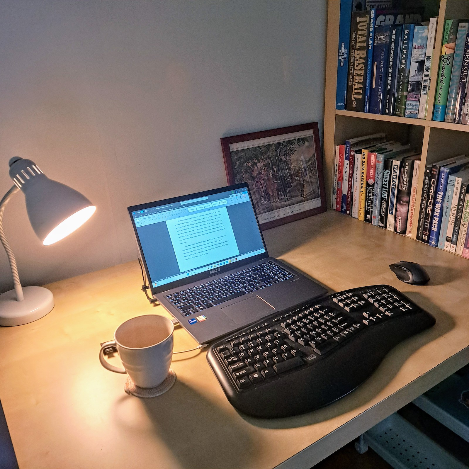 A photo of Andrew Forbes' writing space, showing a laptop with an external keyboard and mouse, a coffee mug, and an illuminated desk lamp. A bookshelf sits at one end of the desk, full of baseball and other books.