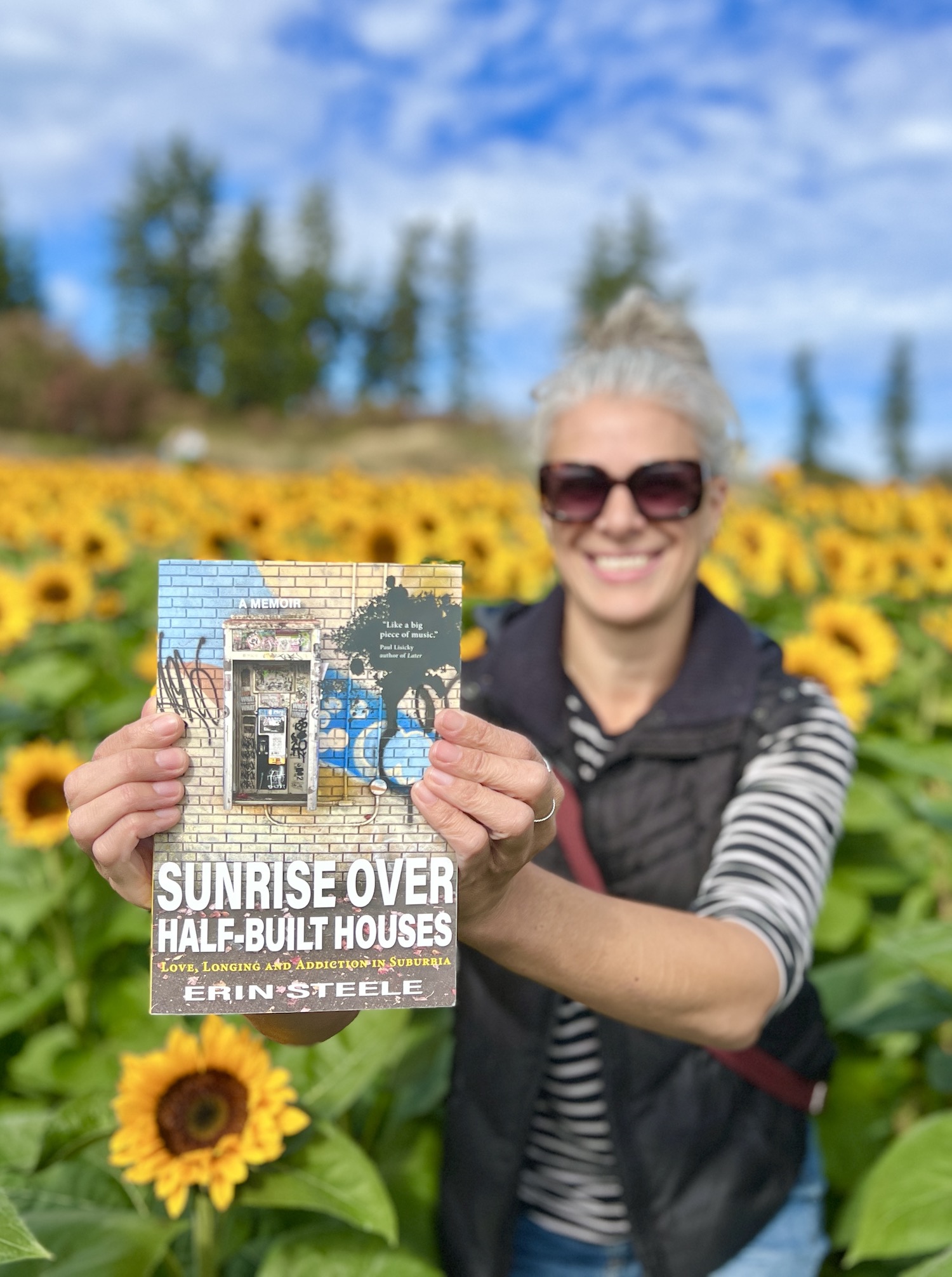 A photo of writer Erin Steele holding up her book, standing in a field of sunflowers.