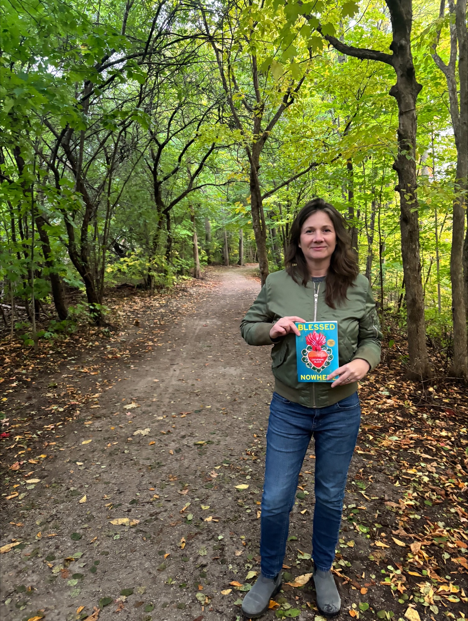 A photo of author Catherine Black, a light-skin-toned woman with brown shoulder-length hair, wearing a green jacket and blue jeans, standing on a nature trail, holding a copy of her book Blessed Nowhere.