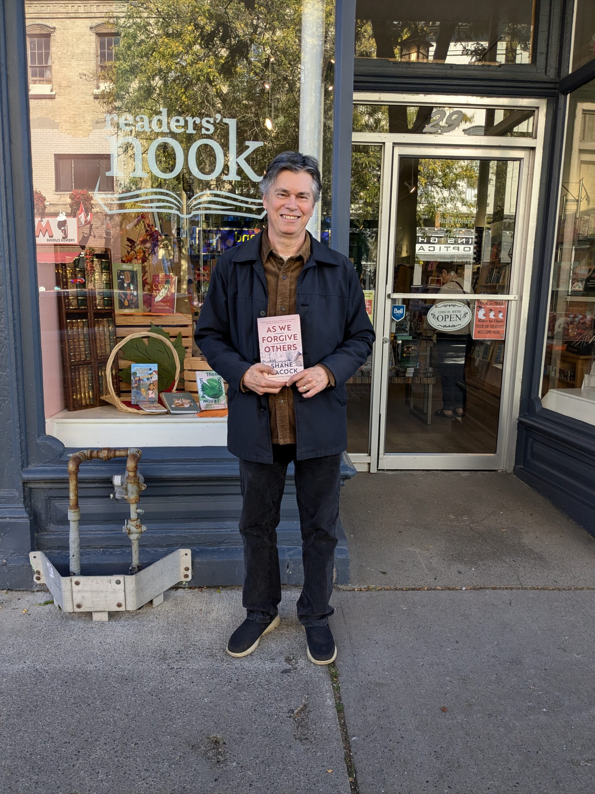 A photo of author Shane Peacock, a light-skin-toned man with short hair wearing black pants and a navy coat and standing in front of Reader's Nook bookstore holding a copy of his book As We Forgive Others.