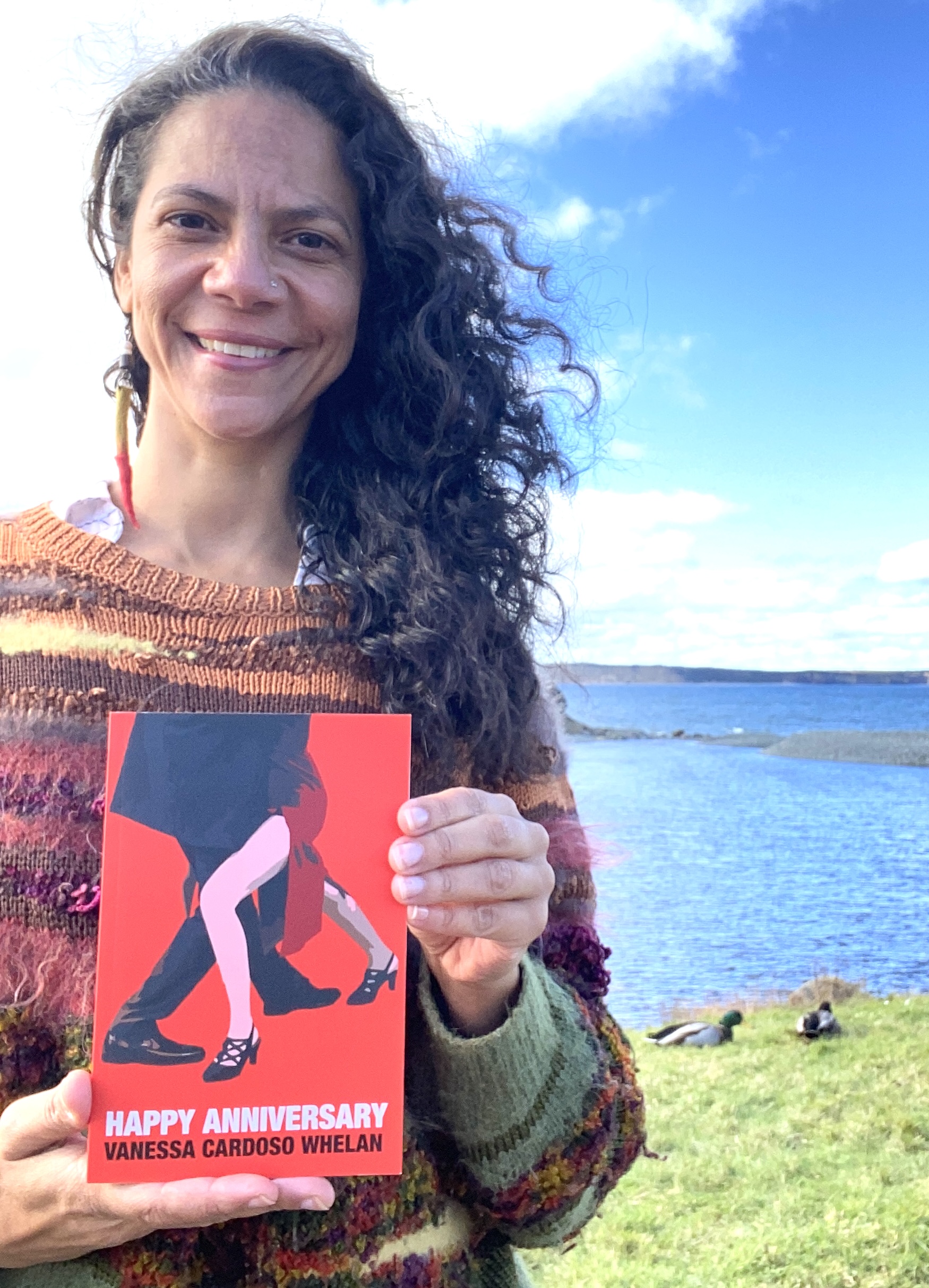 Photo of author Vanessa Cardoso Whelan, a woman with dark curly hair wearing a multi-coloured sweater and standing in front of a body of water. She is holding a copy of her book Happy Anniversary and smiling into the camera. 