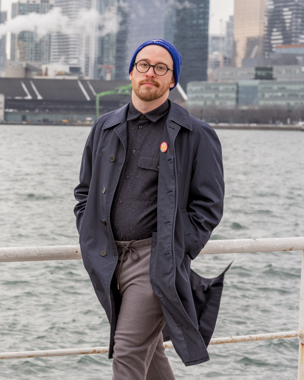 A photo of poet Jake Byrne. They are a light skin-toned person with a light goatee, wearing thick-rimmed glasses and a toque, a long overcoat, and a sweater. They stand in front of choppy waters on an overcast day, with a city skyline beyond the water.