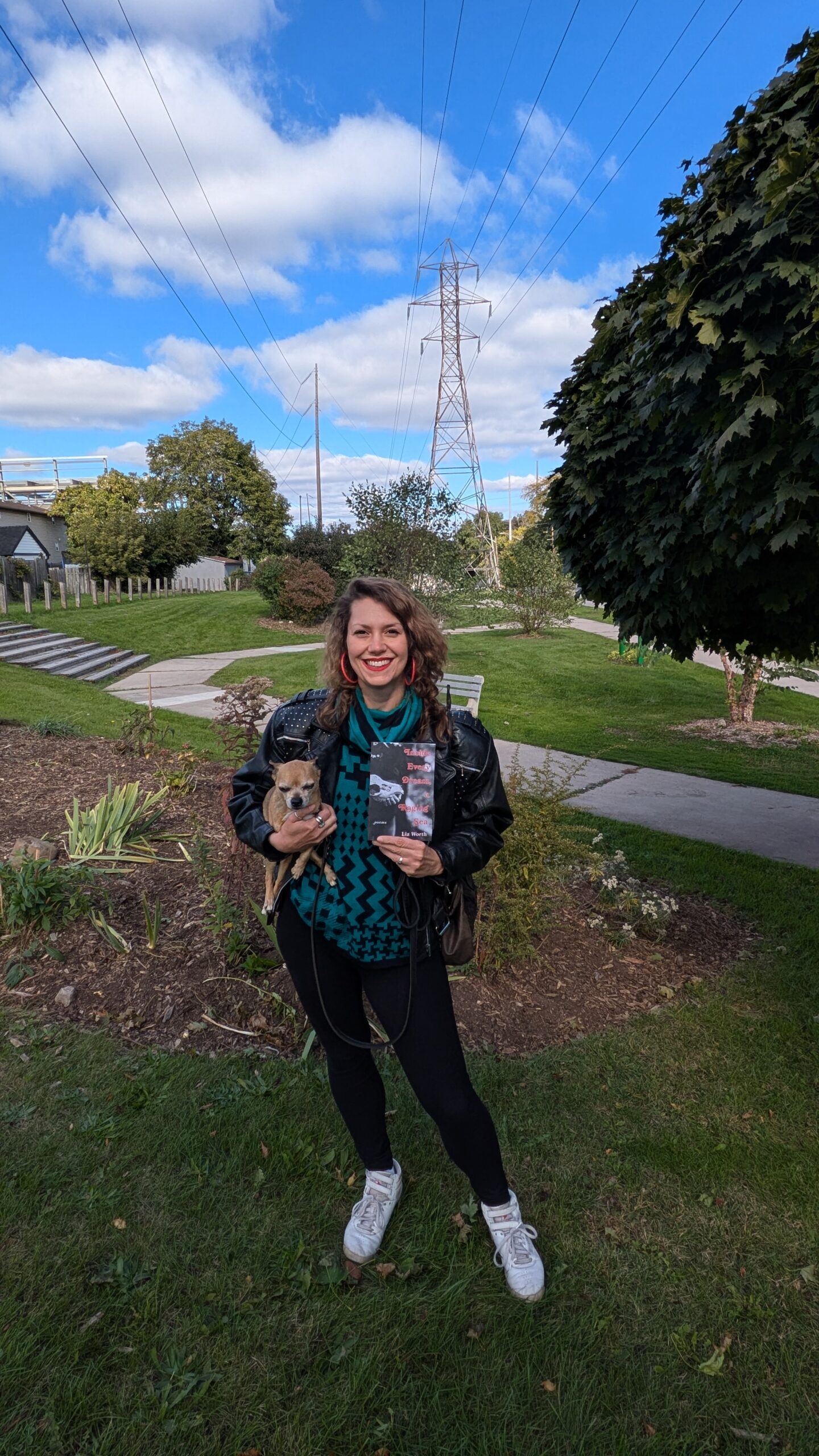 A photo of Liz Worth, a light-skin-toned woman with light-brown wavy hair wearing black pants, a green shirt, a black jacket and pink hoop earrings. She is holding her book Inside Every Dream, a Raging Sea in one hand and a small dog in her other hand and is standing in a parkette in Hamilton, Ontario.