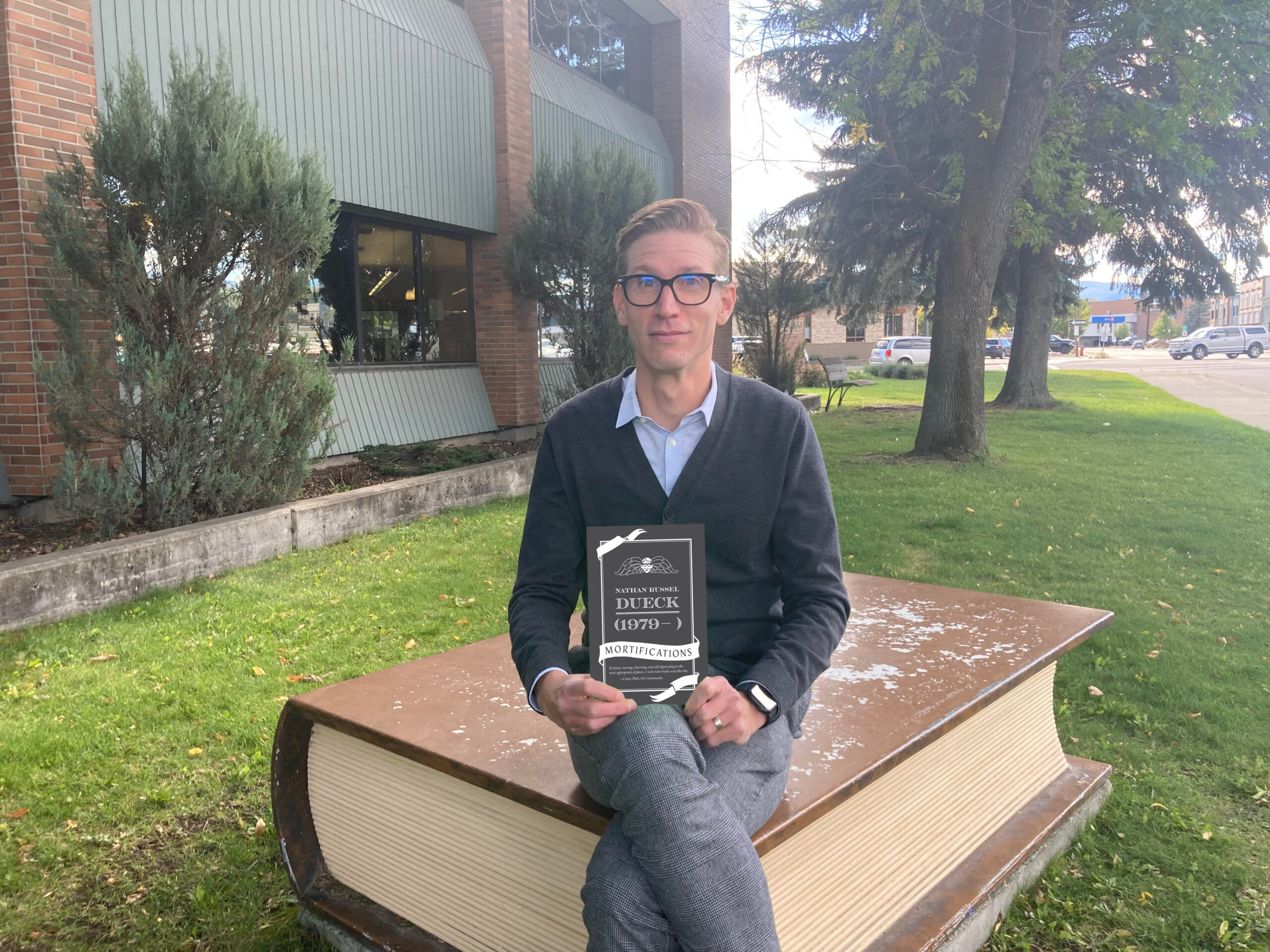 Author Nathan Dueck, a light-skin-toned man with short blonde hair and dark-rimmed glasses wearing a blue collared shirt under a dark-grey sweater and grey pants, sitting on a statue of a book. He is holding his book (1979-     ) Mortifications. 