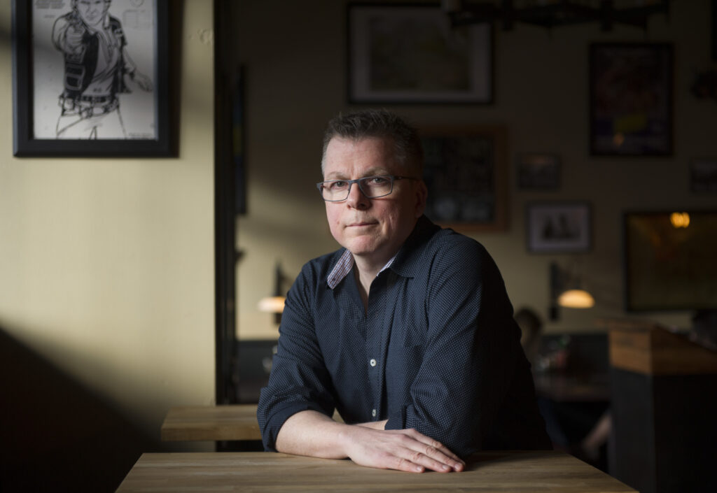 Photo of Peter Darbyshire, a light-skin-toned man with short dark hair and glasses. He is wearing a blue button-up collared shirt and is sitting at a table with his hands resting on the table.