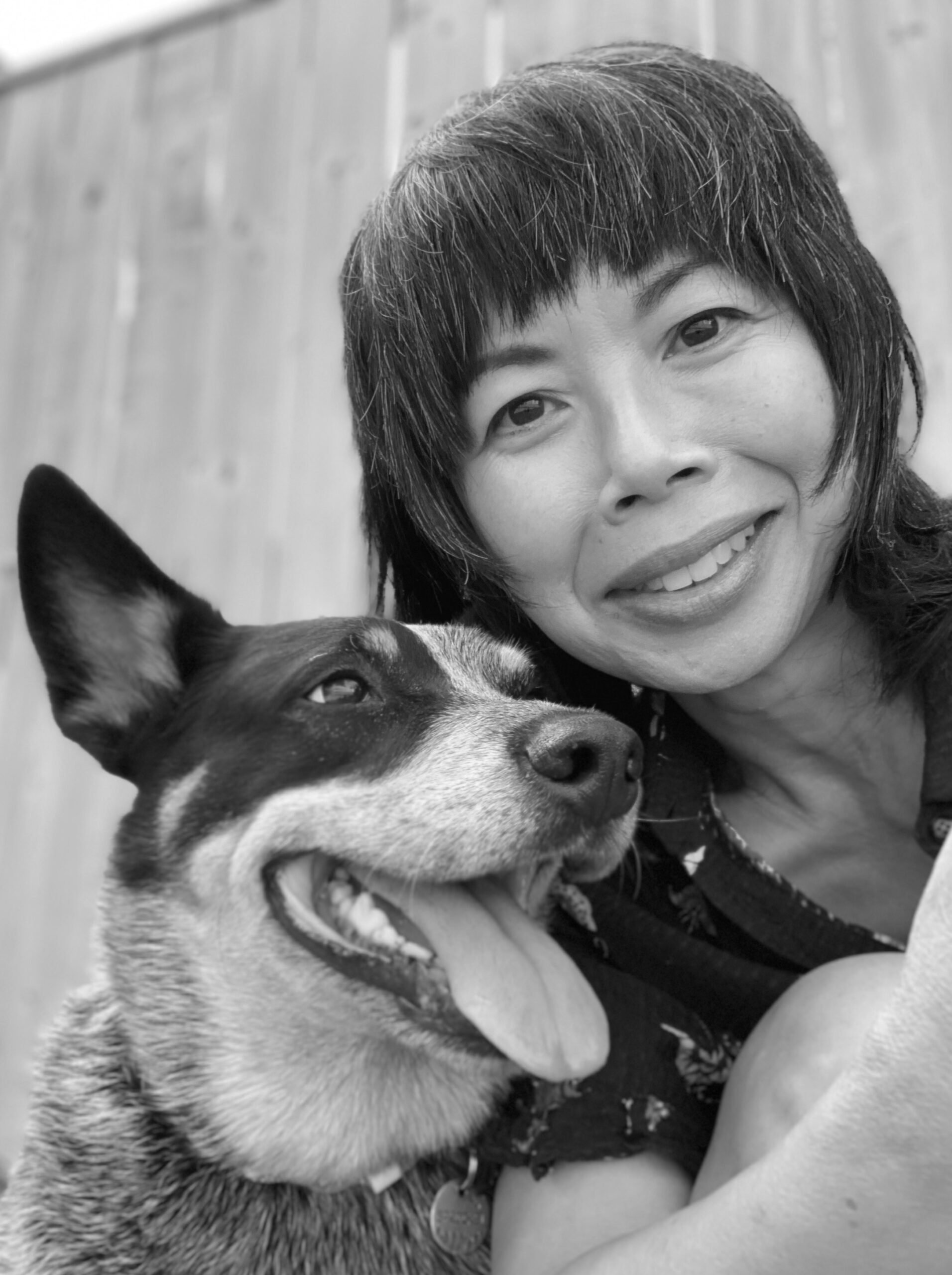 A photo of writer Barbara Tran. She has dark hair cut into bangs and poses with a smiling dog, standing in front of a fence.