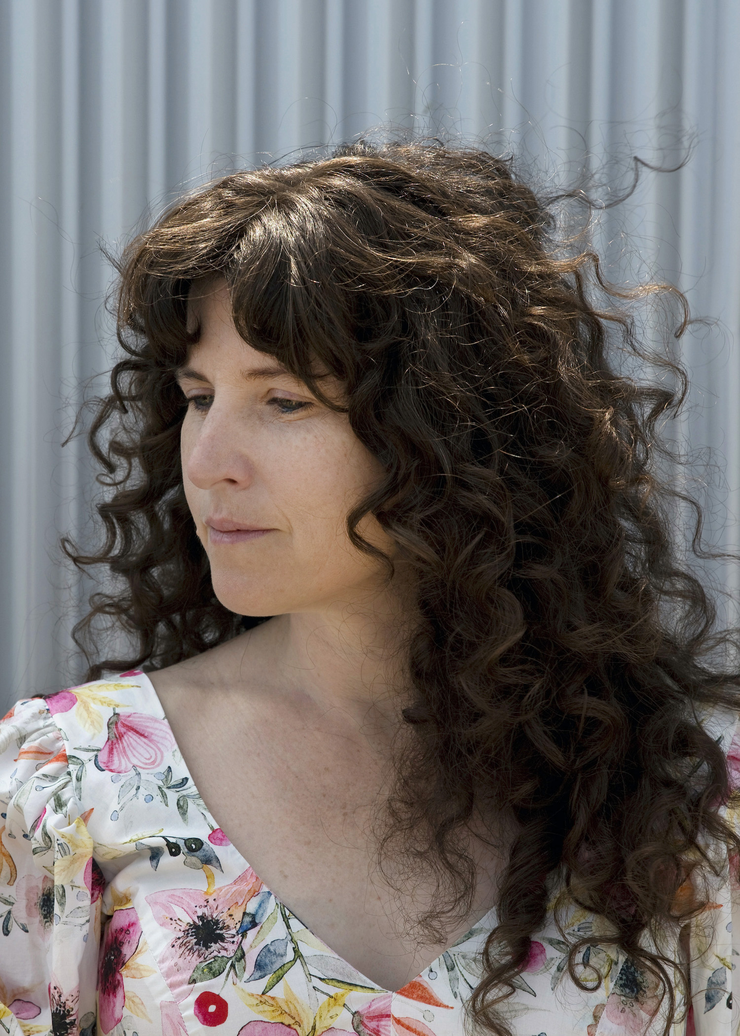 A photo of writer Erin Brubacher. She is a light skin-toned woman with long, dark curly hair. She looks off to the left and wears a brightly patterned top.