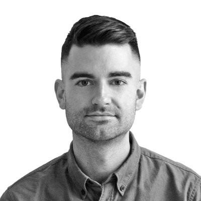 A black and white photo of writer Corey Bialek. He is a light skin-toned man with short, neat dark hair and light stubble, wearing a medium-toned button-down shirt.