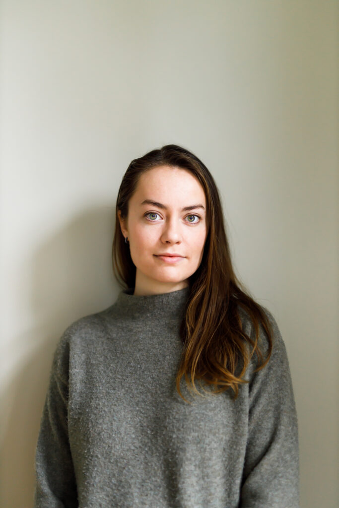 A photo of writer Erica McKeen, who is a light skin toned woman with long brown hair, wearing a grey turtleneck and standing before a light coloured wall.