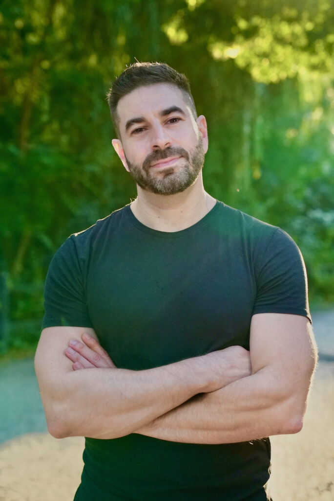 A photo of Matthew Fox. He is a light skin toned man with dark, short hair and a dark, short beard, wearing a dark green t-shirt with his arms crossed over his chest. He stands in a sunny forest.