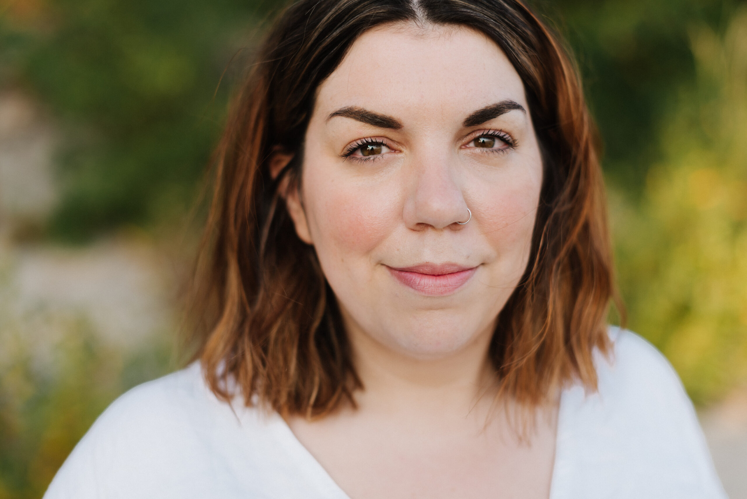 A photo of author Amanda Merpaw, a light-skin-toned woman with short auburn hair and a nose piercing. She is outdoors and the background is blurred.