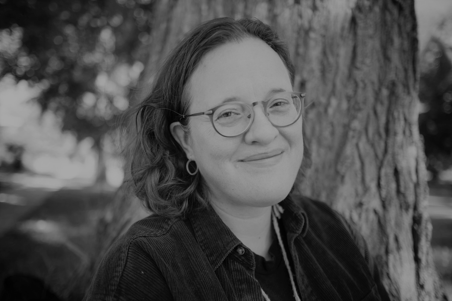 A black and white photo of Ariel Gordon. She is a light skin-toned person with chin-length hair and glasses, and stands in front of a tree outside.
