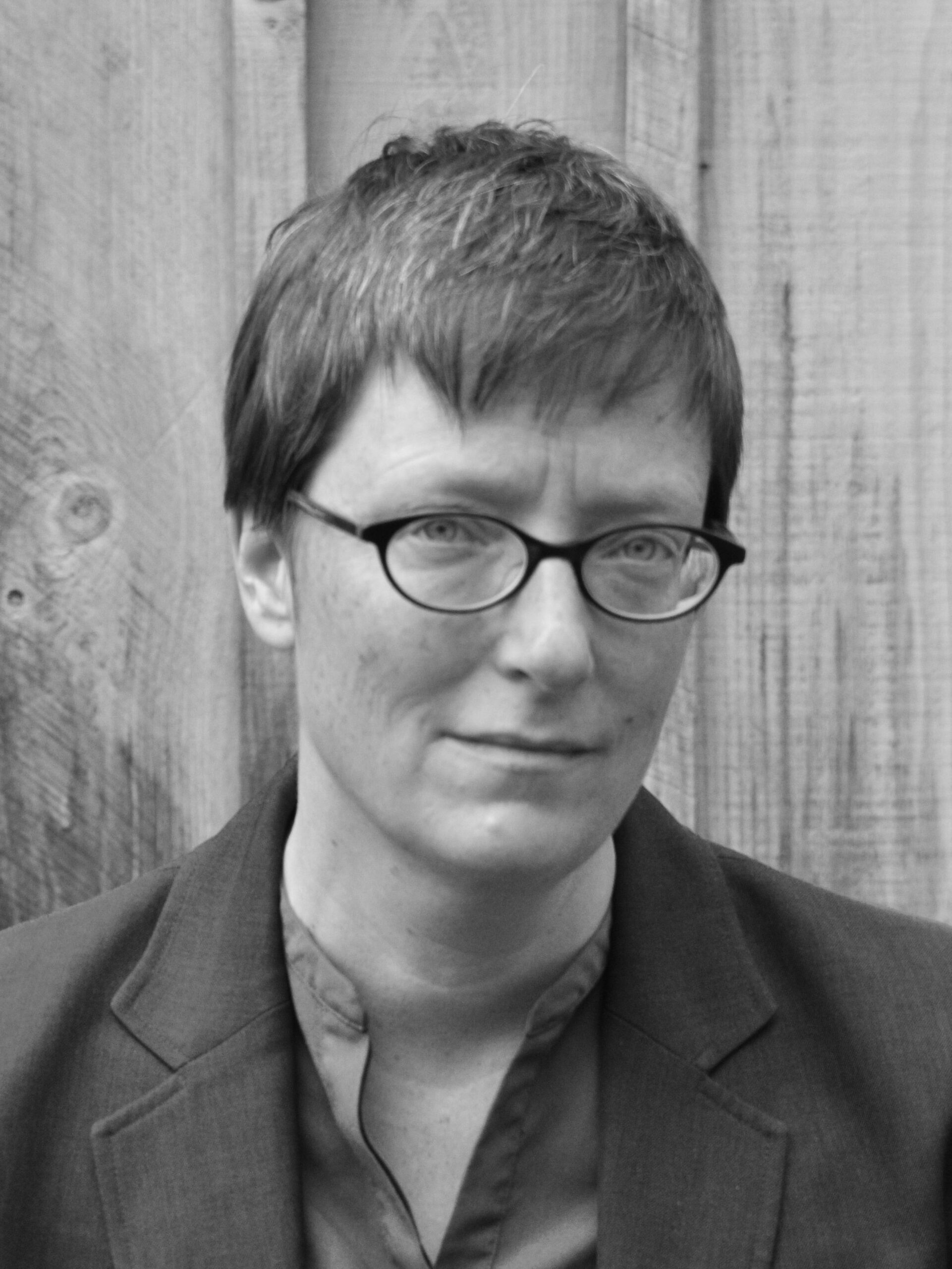 A black-and-white photo of Kate Cayley, a woman with short hair and glasses standing outdoors against a wooden fence. 