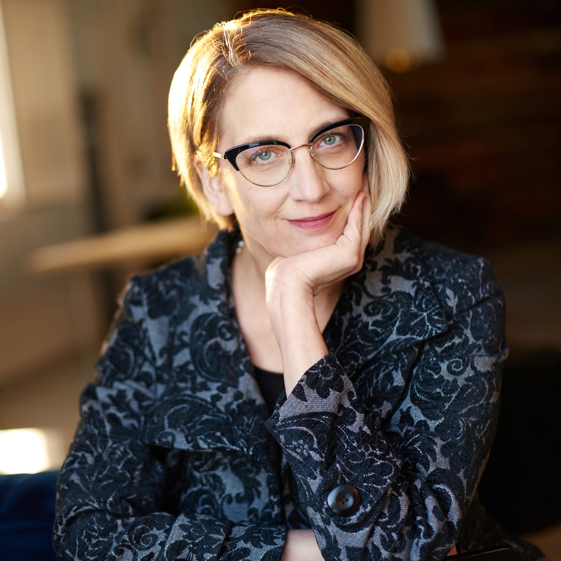 A photo of children's author Leanne Shirtliffe. She has blonde hair cut into a bob, wearing glasses and a blue printed blouse. She rests her chin on her palm.