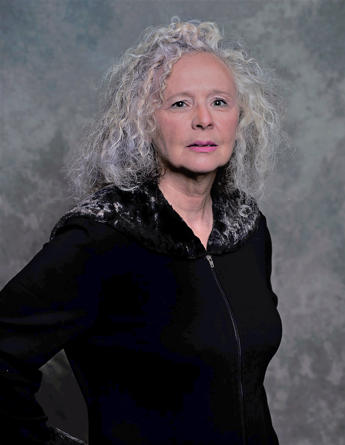 A photo of writer Elaine McCluskey. She is a light skin-toned woman with grey curly hair that reaches her shoulders, and wears a black zip sweater with dark fur on the collar, standing against a grey backdrop.