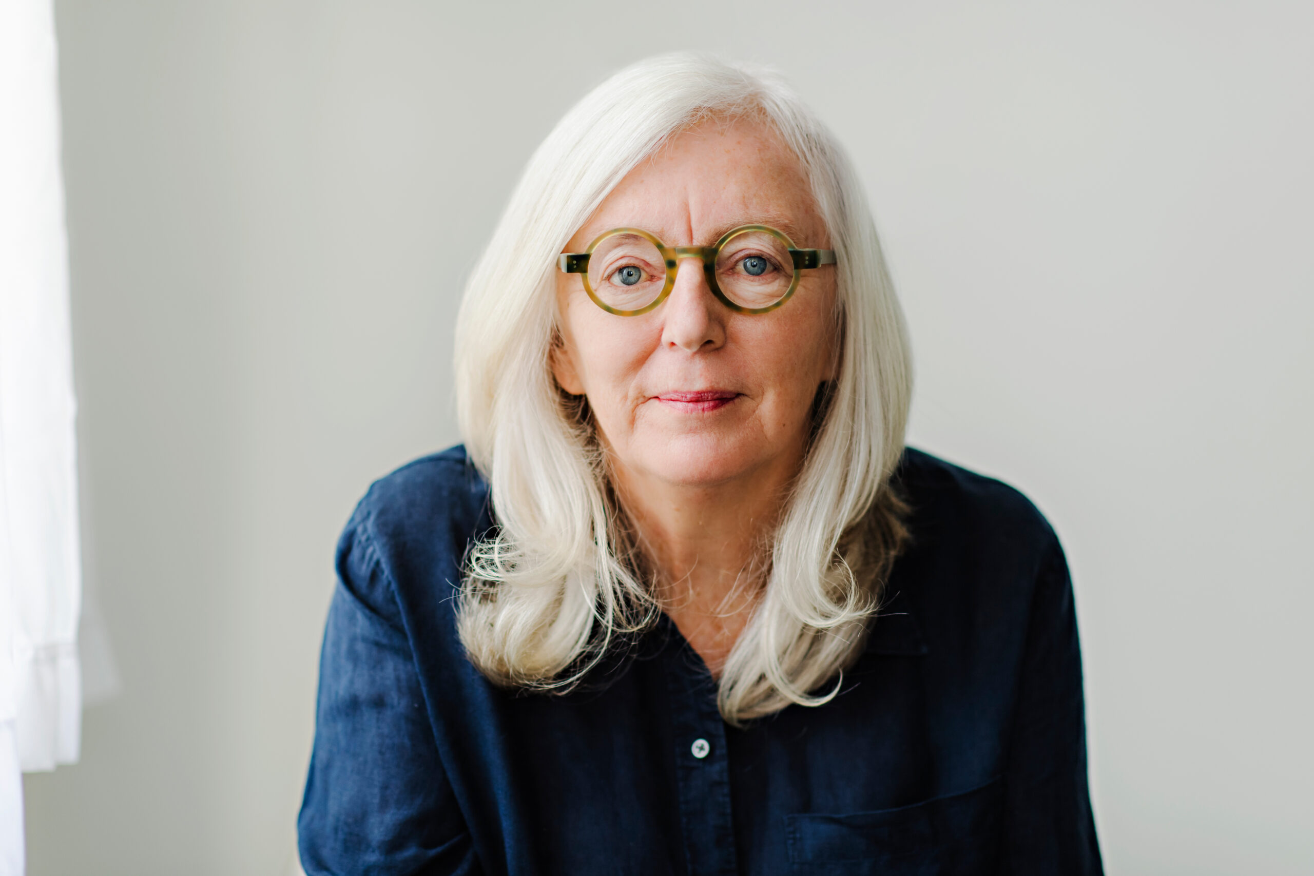 A photo of Pamela Mulloy, a light-skin-toned woman with shoulder-length white-blond hair and glasses. She is wearing a navy-blue button-up shirt and is sitting against a grey wall looking into the camera. 