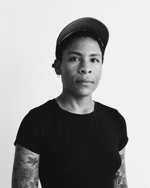 A photo of writer Valérie Bah. They are a nonbinary, Black person with close-cropped dark hair, wearing a baseball cap, nose ring, and black t-shirt. They have floral tattoos on their arms.