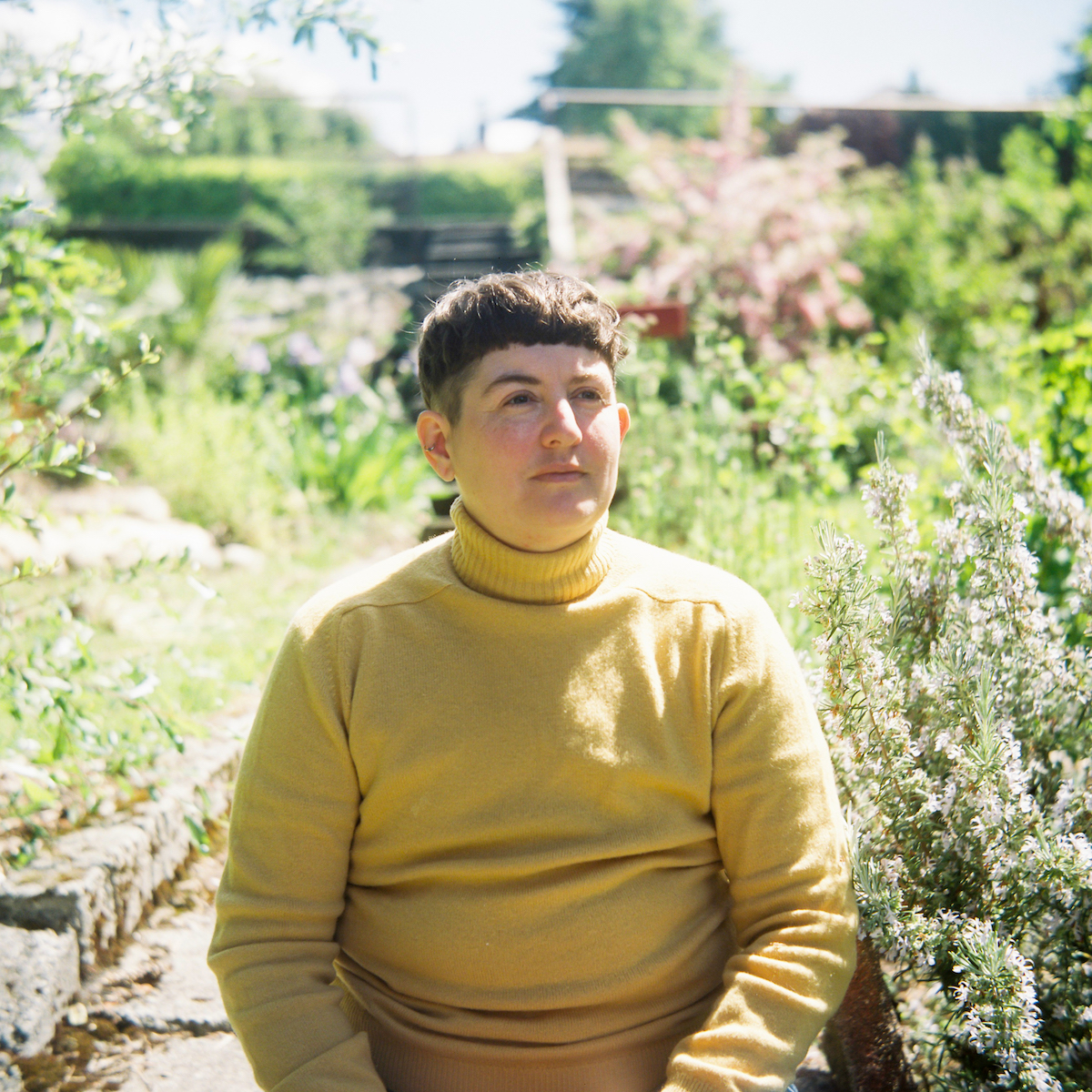 A photo of writer andrea bennett, standing out in a garden of tall, conical flowers. They are a non-binary, light-skin toned person with short dark hair, wearing a yellow turtleneck, looking up towards the sunshine.