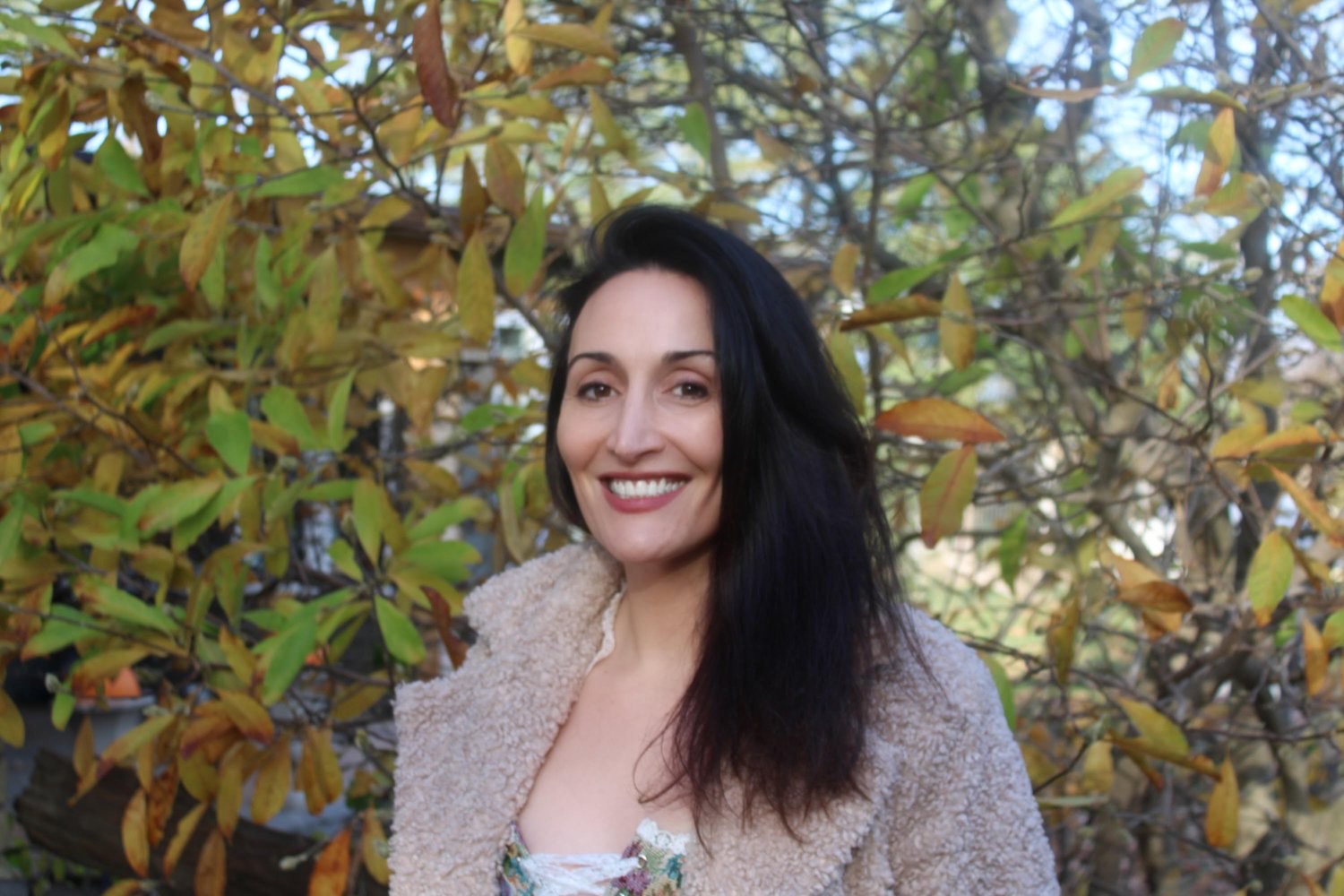 A photo of author Hollay Ghadery. She stands outside, among a tall leafy plant, and smiles at the camera, her dark hair hanging over one shoulder.