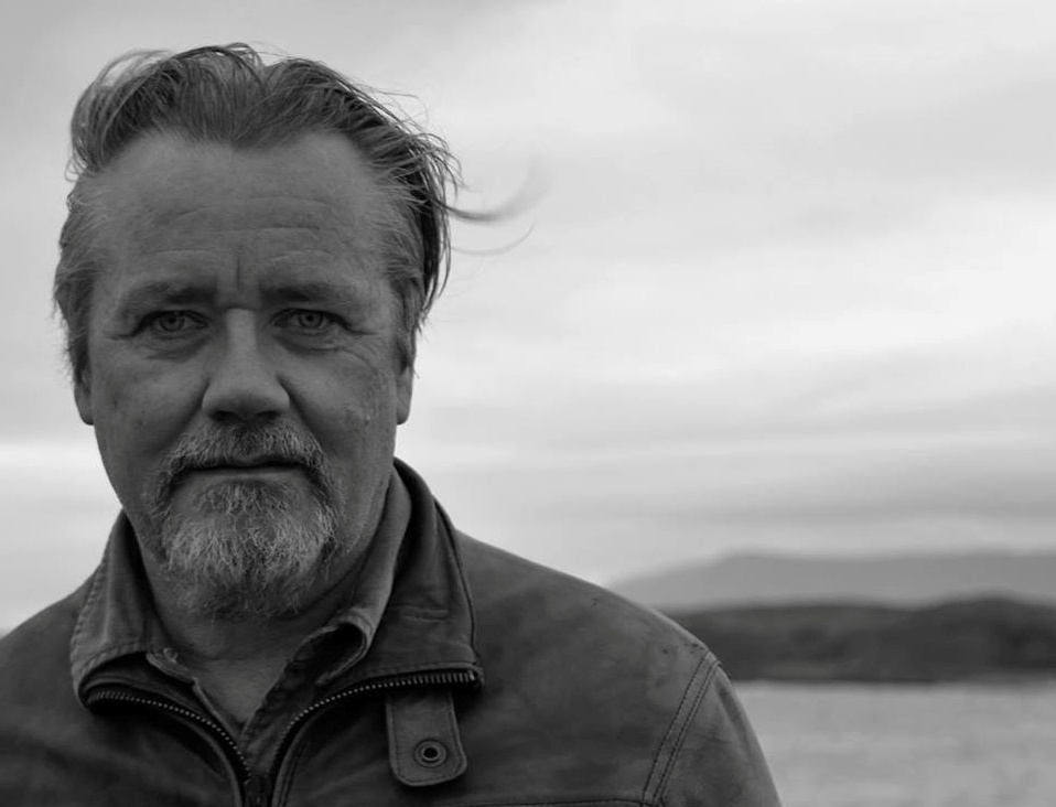 A black-and-white photo of author Gerard Beirne. He is a light-skin-toned man with facial hair. He is standing by a body of water with hills in the background. 