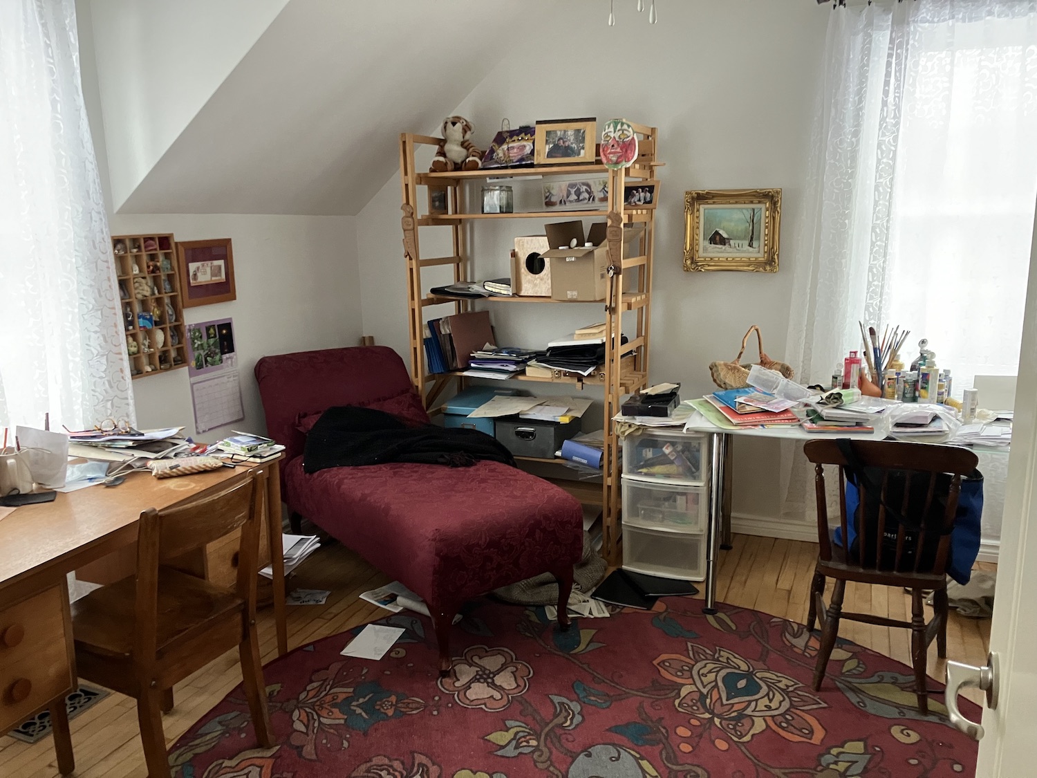 A photo of Margaret Macpherson's workspace. It is a brightly lit room at the top of a house with a slanted ceiling and windows on at least two sides. Two desks piled with printed material, a bookcase, and a burgundy chaise longue fill the space. A printed rug and a little curio shelf add to the uniqueness of the space.