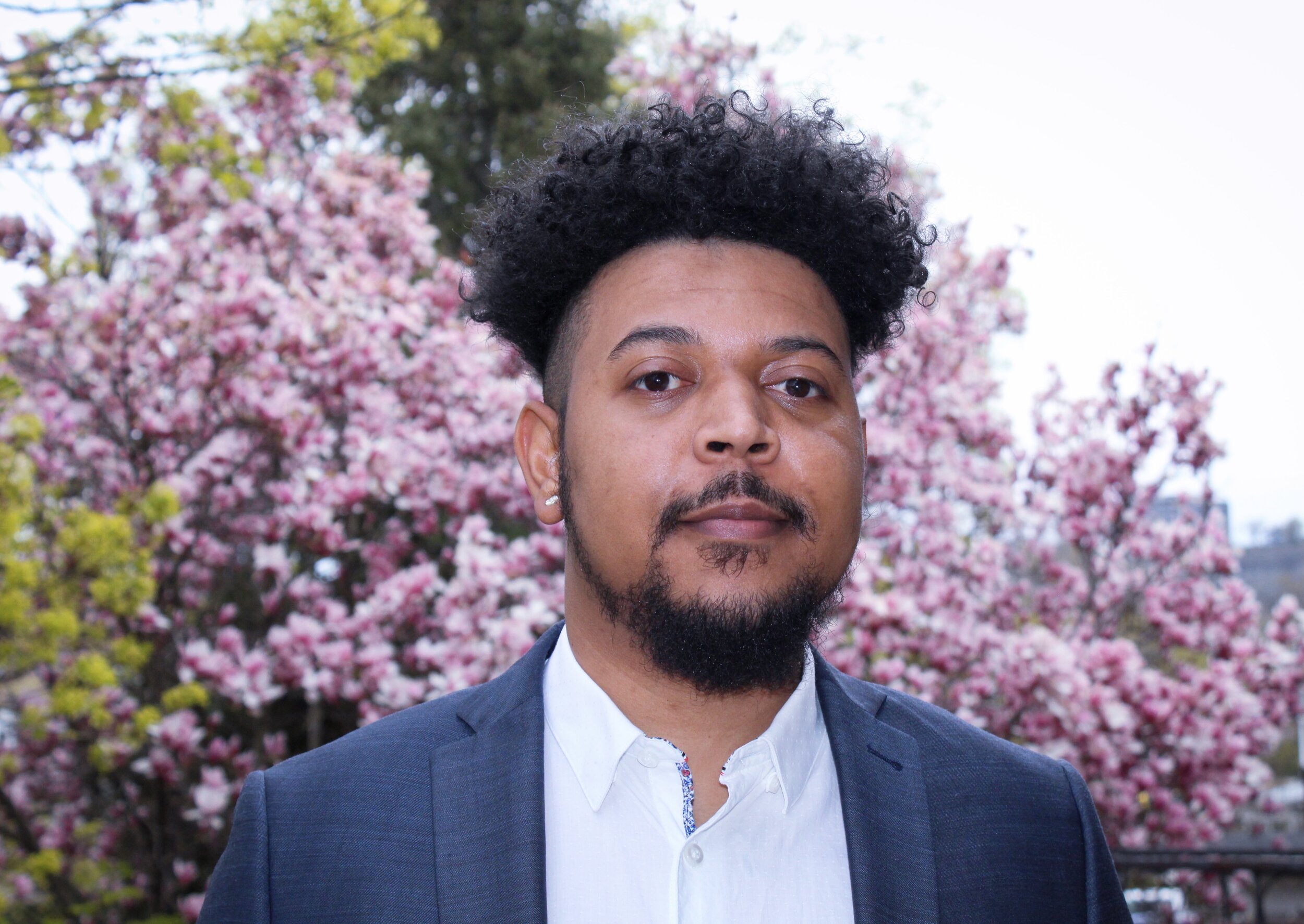 A photo of poet Linzey Corridon. He is a West Indian man with dark Jheri curls and a fade, a mustache and a goatee. He wears a grey suit jacket and a white collared shirt and stands outside, with a magnolia tree in full bloom behind him.