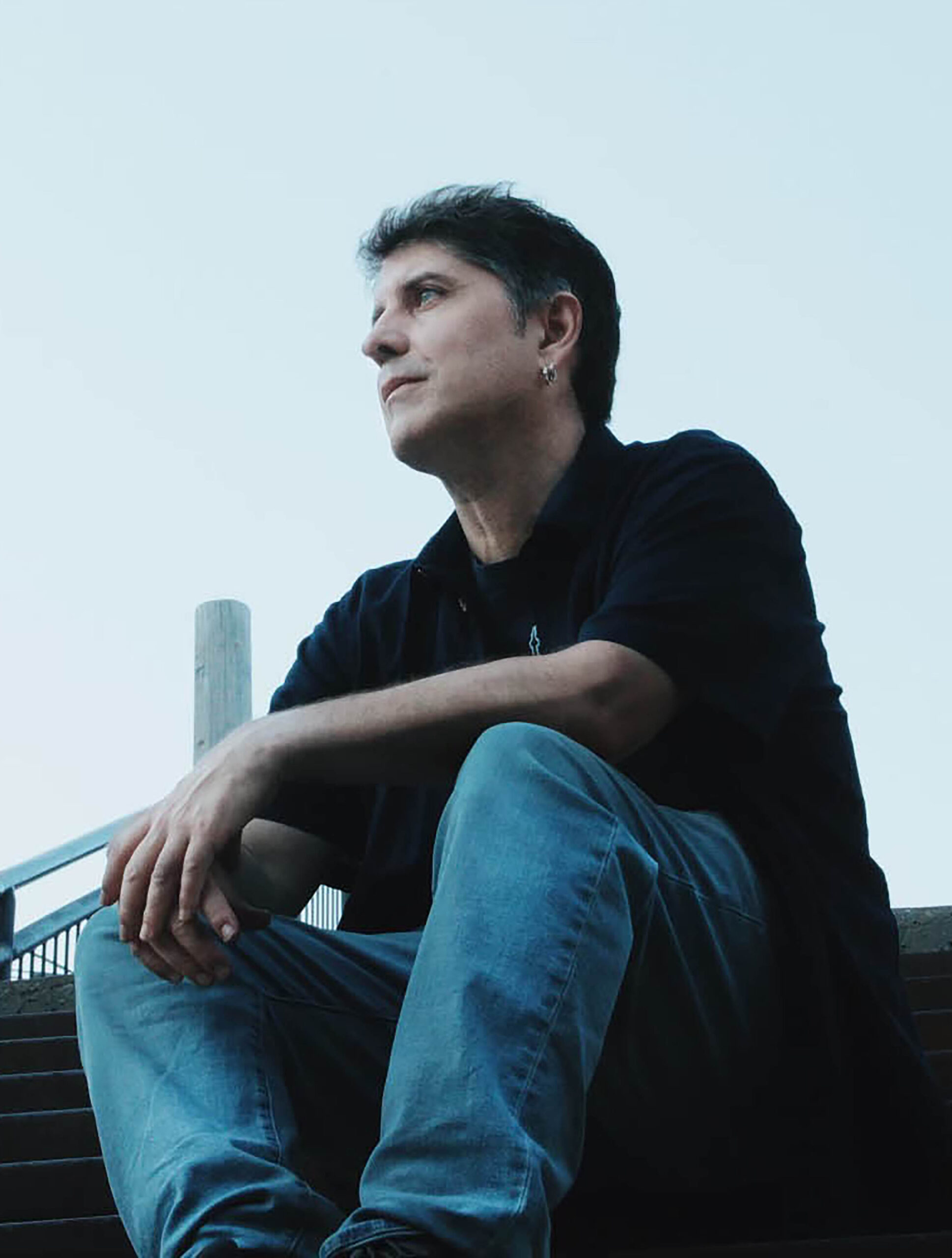 A long-angle photo of Stephen Cain. He has short dark hair and wears a black top and blue jeans. He is sitting outdoors on some steps. 