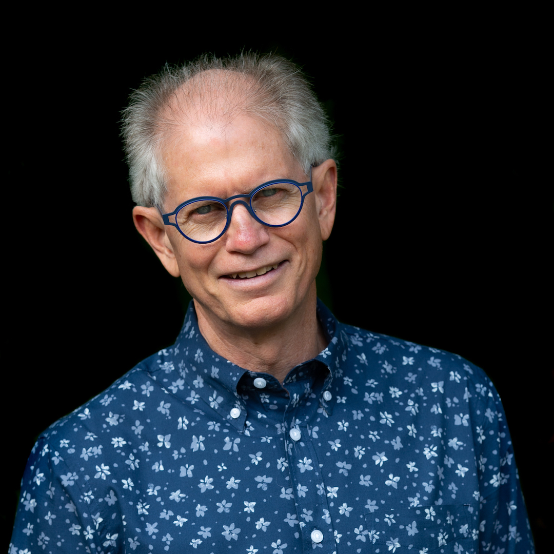 A photo of writer Daniel Coleman. He is a light skin-toned man with grey hair at the sides of his head, wearing bright blue-framed glasses and a blue patterned shirt.