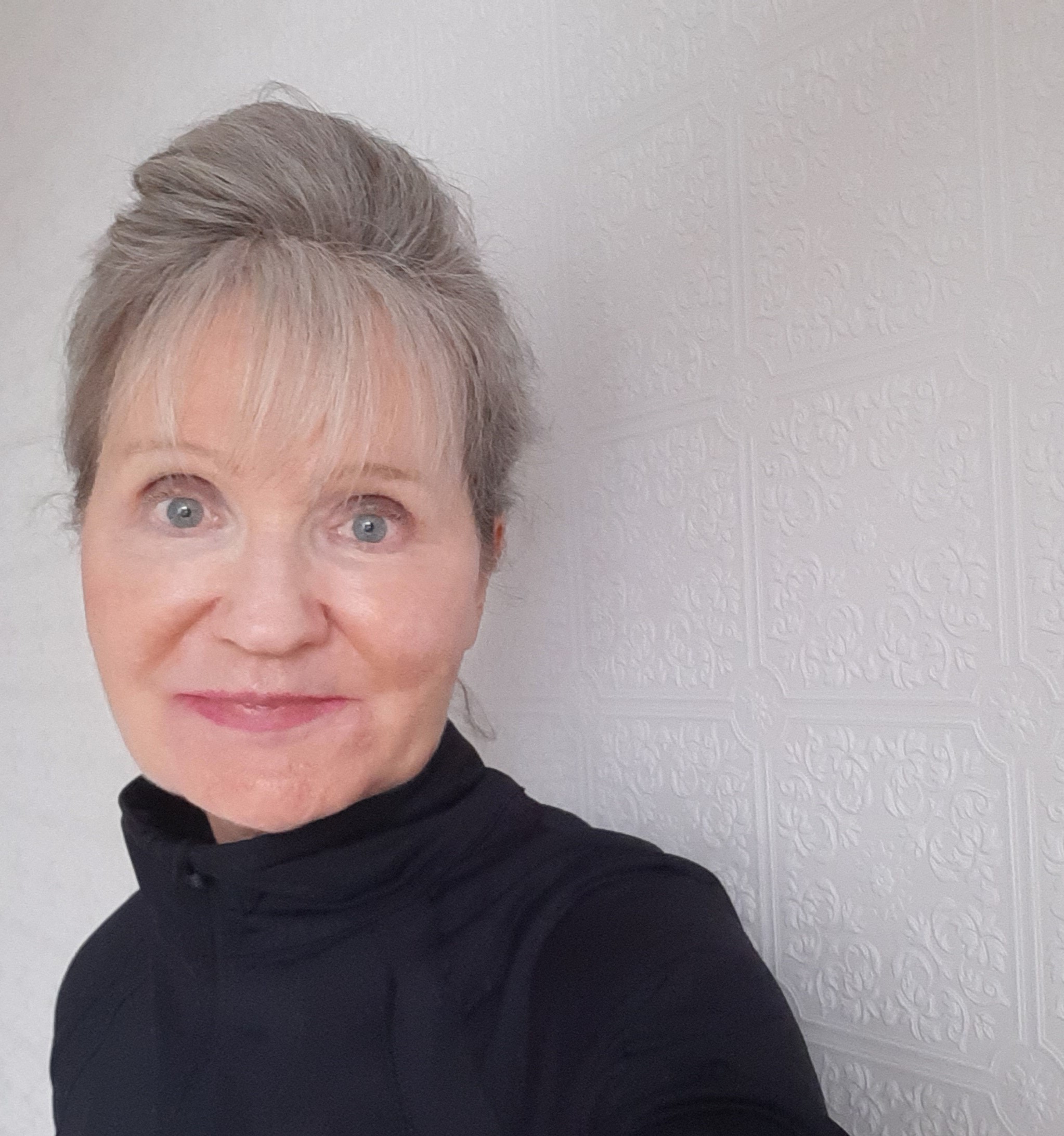 A photo of Elizabeth Murphy. She is a light skin-toned woman with greyish hair and blue eyes. She is wearing a black turtleneck and standing against a white wall, looking into the camera at an angle. 