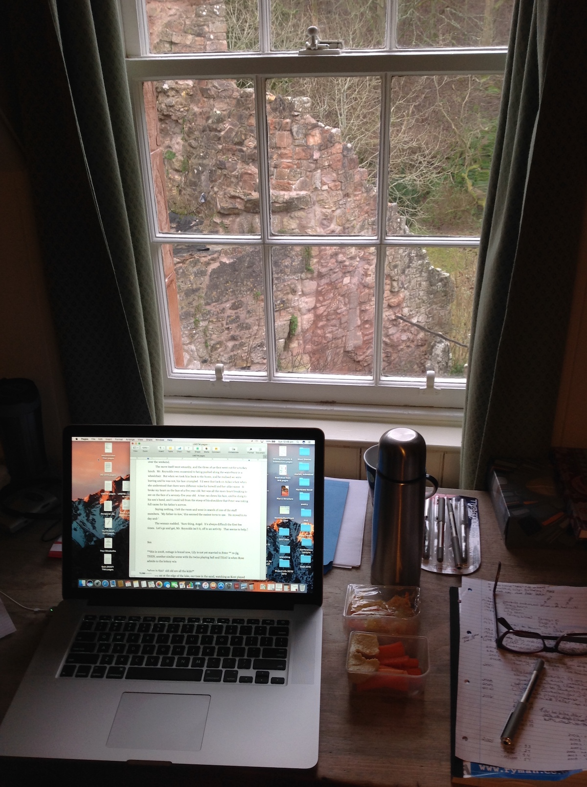 An open laptop on a wooden table with various writing instruments: a pair of glasses, a snack, a coffee thermos. The window view beyond reveals a crumbling, red stone wall.
