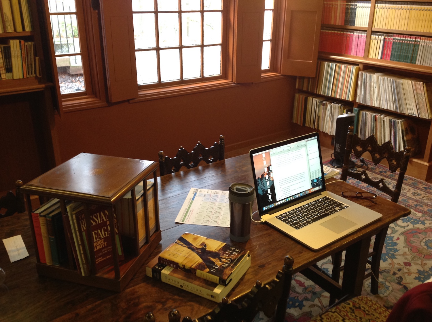 A photo of Louise's writing space at Hawthornden Castle. A laptop sits open on a glossy wooden table overlooking a window. There is a stuffed bookcase against the far wall.